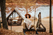 © Алина Троева - Portrait of beautiful brunette woman sitting near the lake, wooden arbor and stairway. Fashion outdoor shot of pleased lady in cozy clothes and straw hat enjoying nature views in autumn countryside.