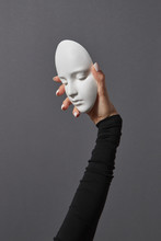 White Plaster Mask Face Is Holding Woman's Fingers. Girl Hand In A Black Sweater On A Gray Background. Concept Social Psychological Masks