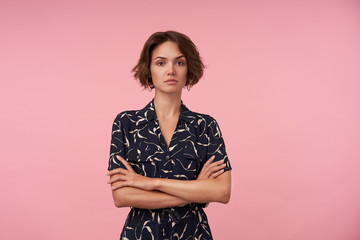 Wall Mural - Portrait of serious young woman with short haircut wearing black romantic dress while standing over pink background, folding hands on her chest and looking at camera with raised eyebrow