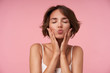 © timtimphoto - Close-up of pleasant looking brunette female with short haircut posing over pink background with eyes closed, holding her face with raised hands and folding lips in kiss