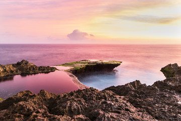 Wall Mural - Devil's Tear at sunset, island of Nusa Lembongan, Bali, Indonesia. Rocky shore in foreground. Tidepool and ocean pink from setting sun's reflection. Pink and yellow sky beyond. 