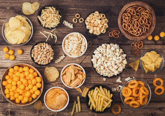 All classic potato snacks with peanuts, popcorn and onion rings and salted pretzels in bowl plates on wooden background. Twirls with sticks and potato chips and crisps with nachos and cheese balls.