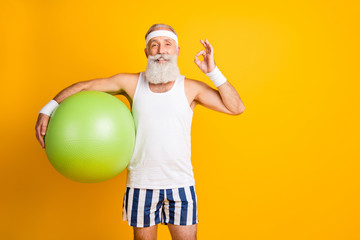 Photo of aged model white hair guy hold green fit ball showing okey symbol advice great trainer wear tank-top striped shorts sweatband isolated yellow color background