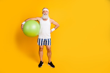 Full body photo of aged senior guy holding big green fit ball ready start training active way of life wear sports tank-top shorts sweatband sneakers isolated yellow color background