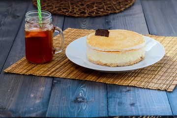 On a dark wooden tabletop on a straw napkin on a round white dish, cake cheesecake and tea with lemon in a glass mug with a green straw.