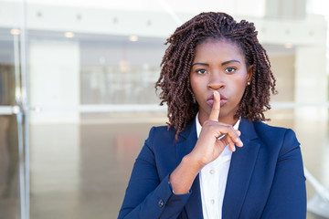 Wall Mural - Serious silent professional keeping secret. Young black business woman standing at outdoor glass wall, applying index finger at mouth, making shh gesture. Conspiracy concept