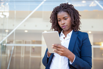 Focused professional using app on tablet outside. Young African American business woman holding digital device, looking at screen, smiling. Online app concept