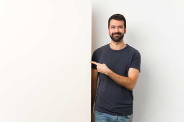 Wall Mural - Young handsome man with beard holding a big empty placard pointing to the side to present a product