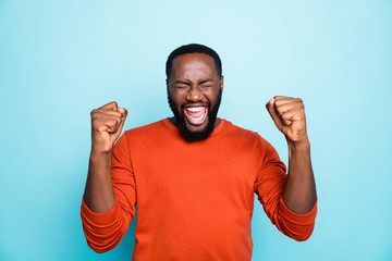 Photo of attractive dark skin guy celebrating lottery winning raising fists excited wear casual orange pullover isolated blue color background