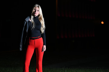 Portrait of a pretty blonde girl at night against the background of the night city. He is standing in front of the camera in a black jacket, against a background of lights.