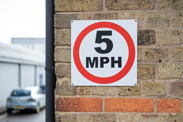 Detail of a 5MPH sign seen attached to a brick wall. The sign, located in a small industrial area, is to promote safety. A car can be seen parked in the distance.