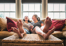 Two Teenage Girls With Phone Sitting On Couch With Feet Up Laughing