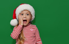 Portrait Of Surprised And Shocked Little Toddler Child Girl In Santa Hat Standing Isolated Over Gree Background. Looking At Camera. Hands Near Open Mouth. Happy New Year And Christmas Holiday Concept	