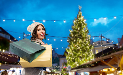 Sticker - seasonal sale and consumerism concept - happy smiling young woman in knitted winter hat and sweater with shopping bags over christmas tree at old town square market in tallinn, estonia background