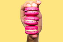 Hand Holding Three Pink Macarons. Close Up Of The Biscuits. Yellow Background. Isolated Subject