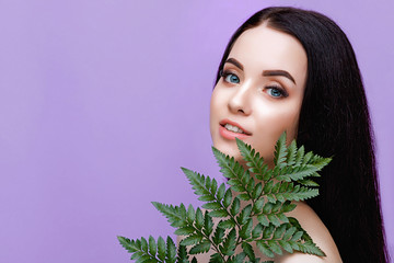 Portrait of young beautiful woman with healthy glow perfect smooth skin and blue eyes holds green tropical leaf. Model with natural nude make up. Purple background.