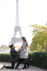 Afro american happy man making proposal to caucasian girlfriend in Paris, Eiffel Tower in background.