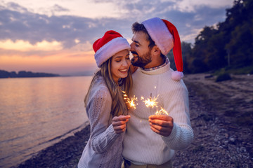 Overjoyed caucasian couple in love standing on coast near river and holding sparklers. Both are dressed in white sweaters and having santa hats on heads.