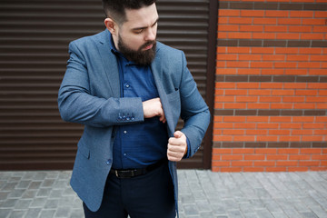 Wall Mural - Stylish handsome brunette man with beard, wearing blue suit, puts a smartphone or wallet in the inner pocket of his jacket against brick street wall. Classic elegant formal men's outfit.