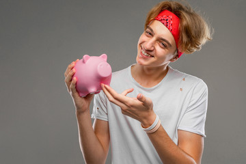 young caucasian man with short red curly hair in white t-shirt, red glasses isolated on a grey background shows a pig moneybox