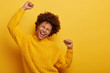 Curly haired girl in winter yellow sweater dances with arms spreading in air, enjoys music, has overjoyed face expression, being on party, enjoys concert poses indoor. People, fun, emotions, happiness