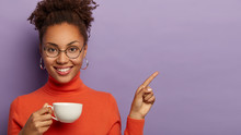 Adorable Curly Haired Woman With Tender Smile, Relaxes With Drink In White Mug, Points On Copy Space, Enjoys Refreshing Coffee, Wears Optical Glasses And Turtleneck, Stands Against Purple Background