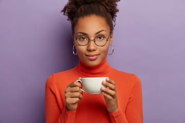 Wall Mural - Serious curly woman in spectacles enjoys hot drink in cold weather, holds white cup of tea, dressed in orange poloneck, looks directly at camera, poses indoor needs morning refreshment drinks espresso