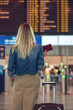 Young blond woman with suitcase and passport at airport against information board getting details about flight.