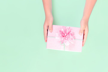 Wall Mural - Hands holding a pink gift box with festive flower on top against pastel green background. Present and celebration or giveaway for feminine bloggers. Mothers Valentines Womens Day