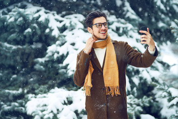 Wall Mural - Young man in a coat on the street with a phone. Smiling young man having fun outdoors. Businessman uses a phone.  Man enjoy a winter. Dressed in a coat, sweater and scarf. Winter concept. Snowfall.