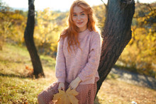 Portraits Of A Charming Red-haired Girl With A Cute Face. Girl Posing In Autumn Park In A Sweater And A Coral-colored Skirt. In The Hands Of A Girl A Yellow Leaf