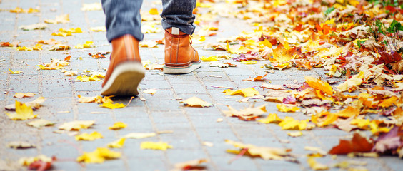 Back view on the feet of a man in brown boots walking along the sidewalk strewn with fallen leaves. The concept of turnover of the seasons of the year. Weather background