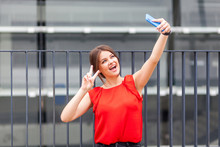 Portrait Of Fancy Young Brunette Woman In Stylish Red Shirt Taking Selfie On Smartphone And Showing Victory Or Peace Gesture, Celebrating Success. Making Video Call, Standing Near Fence Outdoor