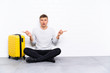 © luismolinero - Young handsome man sitting on the floor with a suitcase pointing to the laterals having doubts