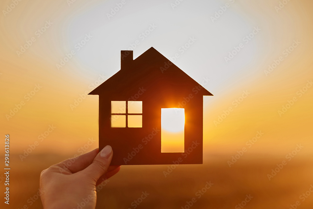 Woman hand holds wooden house against the sun - obrazy, fototapety, plakaty 