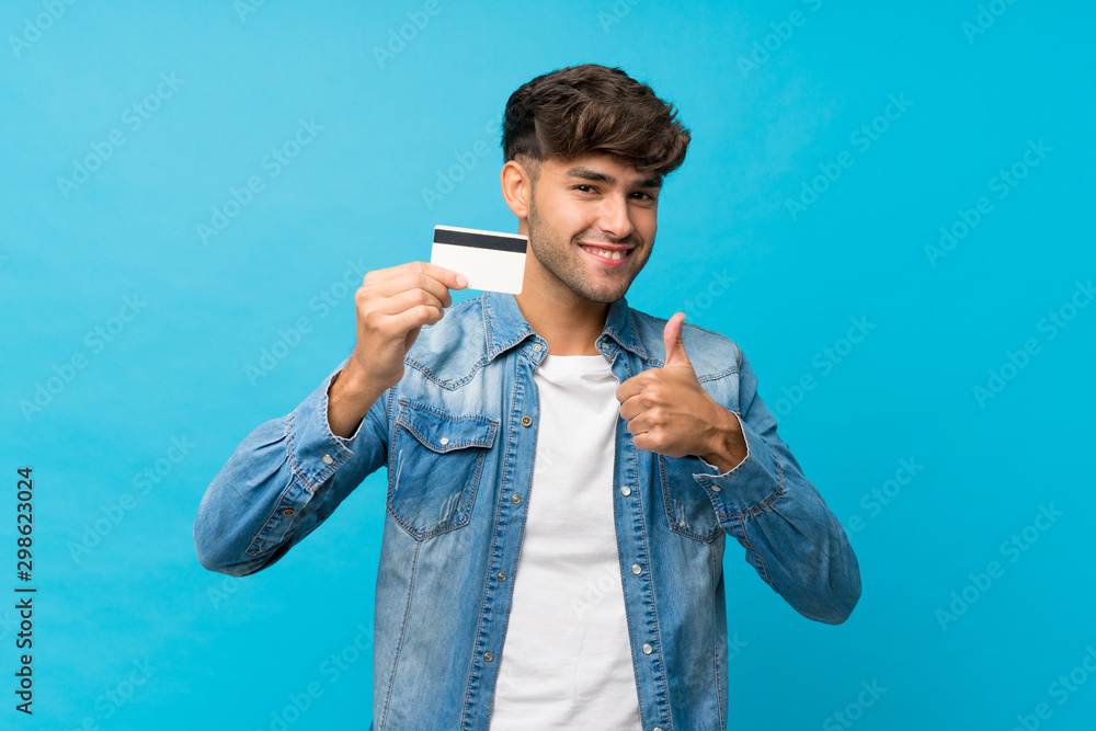 Young handsome man over isolated blue background holding a credit card - obrazy, fototapety, plakaty 