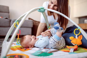 Wall Mural - Side view of happy caucasian mother playing on the floor with her loving baby boy. Living room interior.