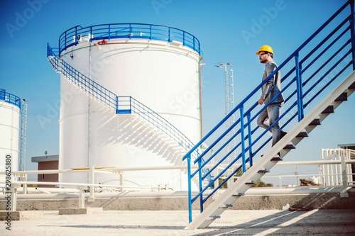 Cuadros en Lienzo Full length of handsome Caucasian businessman in suit and helmet on head going down the stairs on oil tank storage