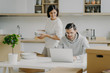 © VK Studio - Young brunette woman unpacks boxes with personal belongings, places plates in cupboard, looks attentively at laptop computer where husband works. Family couple relocate in new modern apartment