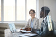 © Seventyfour - Portrait of two young entrepreneurs discussing business project while sitting at meeting table behind glass wall, focus on smiling woman looking at partner, copy space