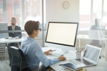 Back view portrait of young office worker using computer with black screen while sitting at workplace, shot from behind glass, copy space