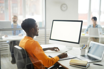 Side view portrait of young African-American man using computer in office, focus on blank white screen, copy space