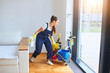 Young caucasian girl with cleaning trolley full of detergents, cleaning room in house with panoramic window. Woman dressed in blue uniform, in process of working