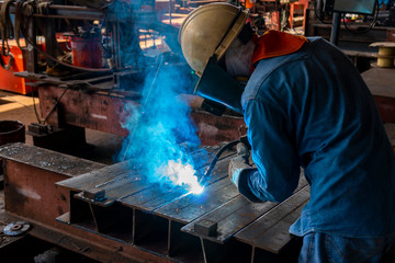 The welder is welding a steel structure with the process Flux Cored Arc Welding (FCAW) in Industrial factory.