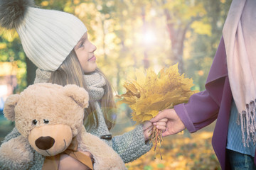 Cute little kid daughter congratulating mum with mothers day, smiling girl with Teddy bear presents autumn yellow leaves, handing them to mother. Bouquet present for birthday, Mopther's Day concept