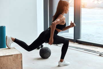 Sportive female in gym doing squats, stretching legs. One leg on fit box. Side view on perfect woman's body