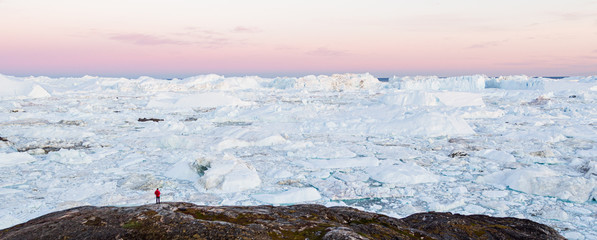 Wall Mural - Global Warming and Climate Change concept. Travel adventure in Arctic landscape nature with icebergs - tourist person looking at view of Greenland icefjord - aerial photo. Man by icebergs, Ilulissat.