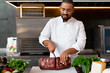 © Andrii - Handsome young African chef standing in professional kitchen in restaurant preparing a meal of meat and cheese vegetables.