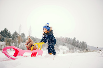 Kid in a winter forest. Girl in a blue hat. Child playing with pink sled