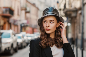 Outdoor close up fashion portrait of young elegant brunette woman wearing faux leather bucket hat, black suit, posing, walking in street of European city. Copy, empty space for text 